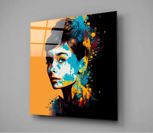 Angled view of glass wall art showing a woman's profile with orange left side, blue and yellow splatter effects on right, mounted on gray wall.