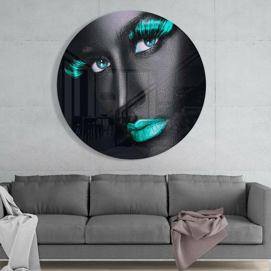 Circular glass wall art depicting a woman's face with gray skin, turquoise eyelashes and lips, displayed above a gray sofa.