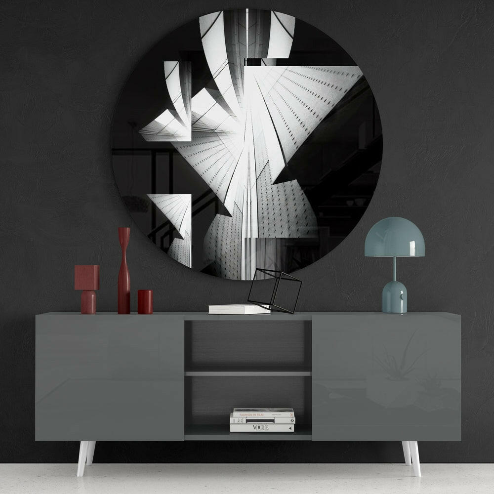 Round black and white glass wall art depicting skyscrapers displayed above gray sideboard with red vases and blue lamp.