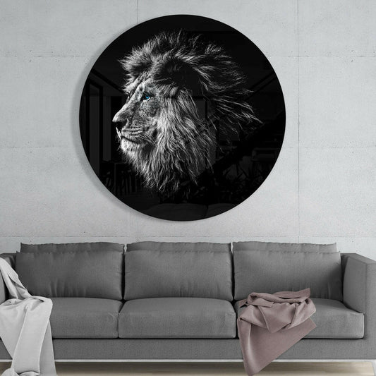 Circular black glass wall art with striking black and white lion portrait displaying piercing blue eyes, mounted above gray sofa in modern interior.