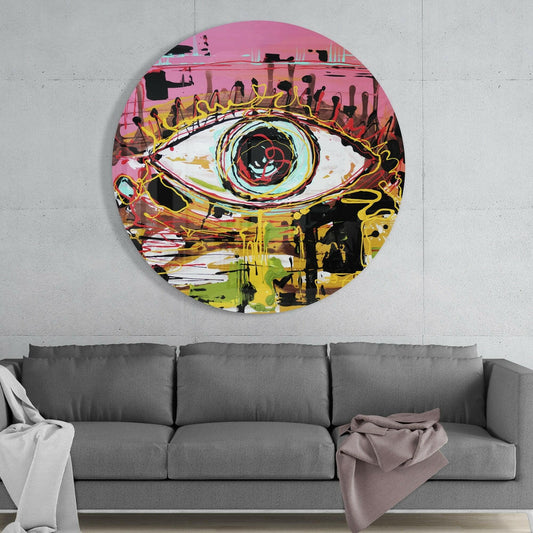 Circular glass wall art with a stylized eye design in pink, yellow, and black hanging above a gray sofa in a living room.