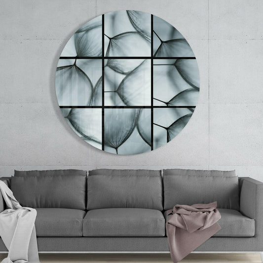 Round glass wall art with black grid overlay displaying grayscale dandelion seed photographs, mounted above gray sofa.