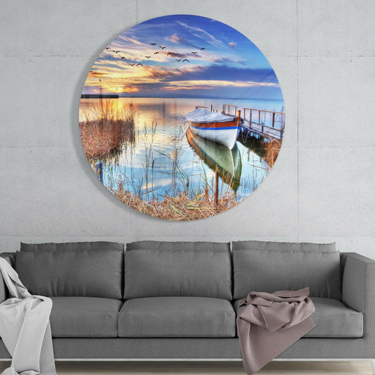 Circular glass wall art mounted above gray sofa displaying sunset lake scene with moored boat, reeds, and flying birds.