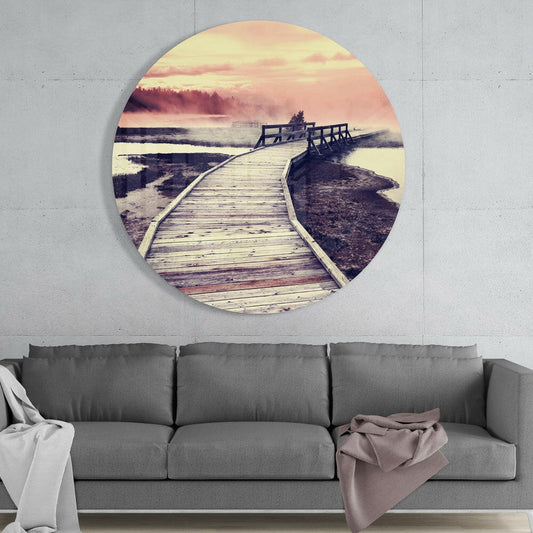 Circular glass wall art mounted above gray sofa showing wooden boardwalk leading toward misty pink sunset over wetlands and forest.