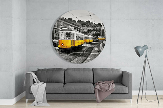 Circular glass wall art mounted above gray sofa showing yellow trams on cobbled street with European buildings, selective color effect against white concrete wall.