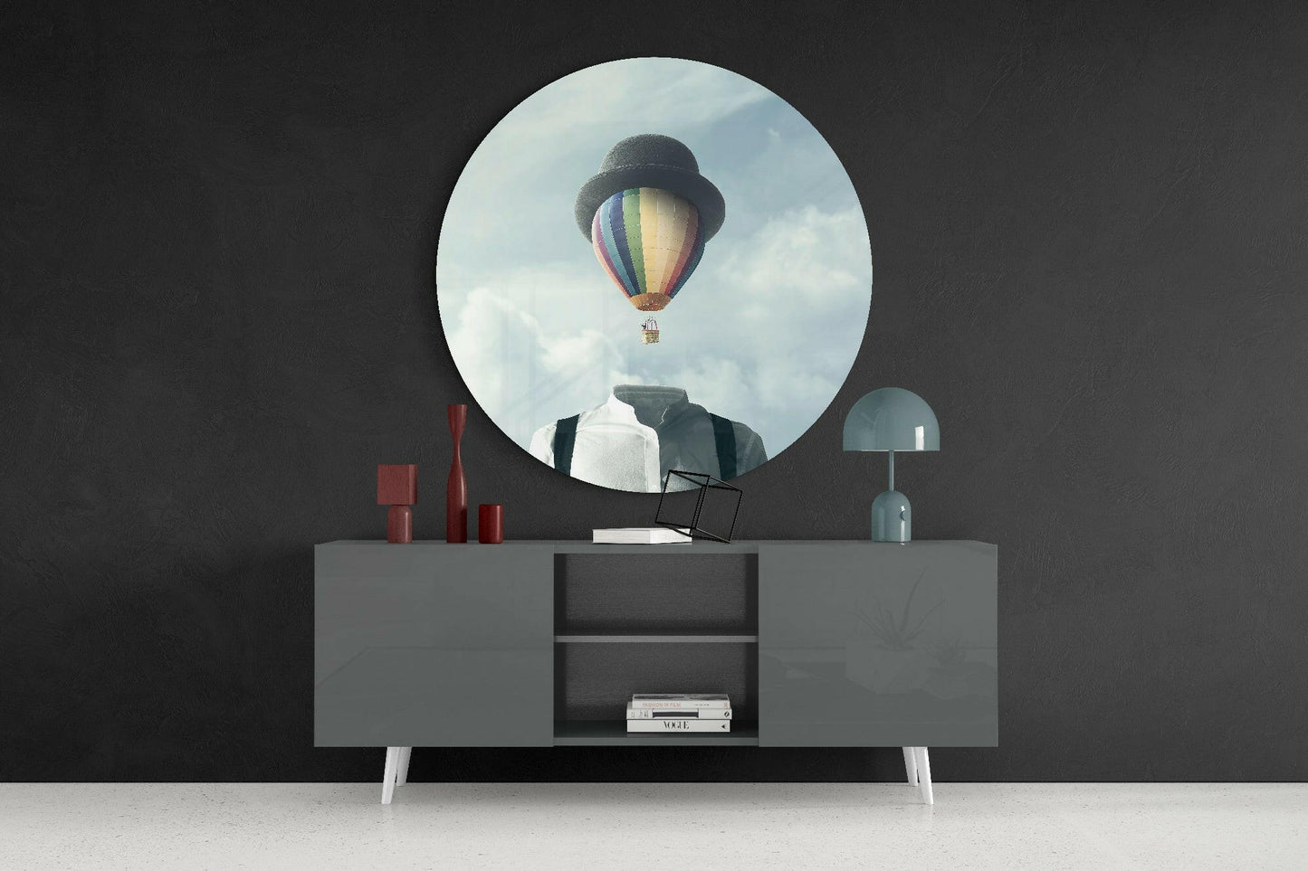 Circular glass wall art depicting colorful hot air balloon in cloudy sky, mounted above gray sideboard with red candles and blue lamp.