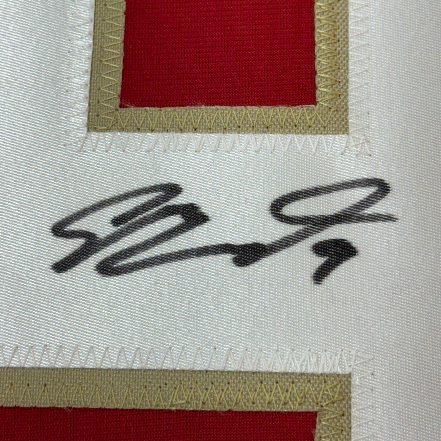 Close-up of handwritten signature "Sam Bennett" in black ink on white fabric with red and tan striped borders.