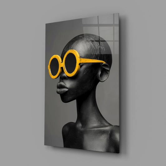 Glass wall art displaying a monochromatic sculptural head in profile wearing oversized yellow round sunglasses against a gray background.