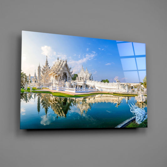 White ornate Thai temple with golden spires reflected in still water, printed on glass panel against gray wall.