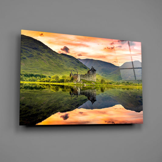 Glass wall art panel displaying a Scottish castle reflected in still water, surrounded by green mountains and golden sunset sky.