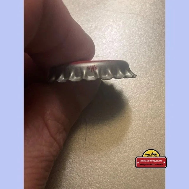 Hand holds a silver crimped metal bottle cap with red lettering on edge.