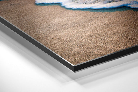 Close-up angled view of brushed aluminium Dibond wall art showing layered edges with sandy beach and turquoise wave imagery.