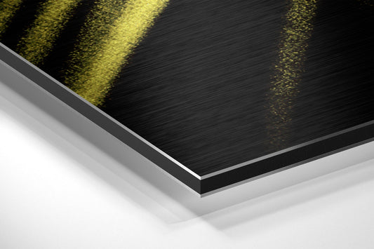 Close-up angled view of layered Dibond panel showing black brushed aluminum surface with yellow brushstroke and white backing layer.