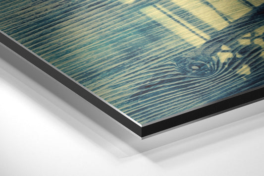 Close-up angled view of layered Dibond wall art showing blue-green brushed aluminum surface with decorative cage pattern and black edge trim.
