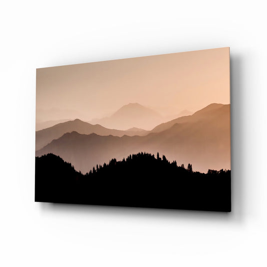Angled view of glass wall art showing sepia-toned layered mountains with silhouetted pine forest foreground against warm peachy sky.