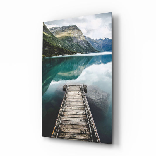 Wooden dock extending into turquoise alpine lake surrounded by forested mountains and cloudy sky, displayed on glass wall art.