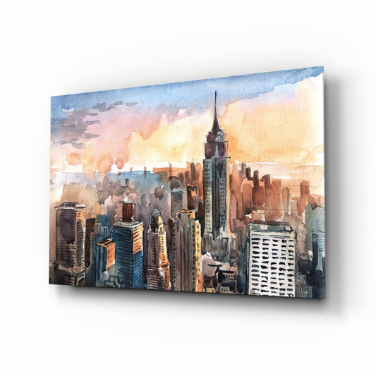 Watercolor glass wall art depicting a Manhattan skyline at sunset with warm orange and blue tones, displayed at an angle against white background.