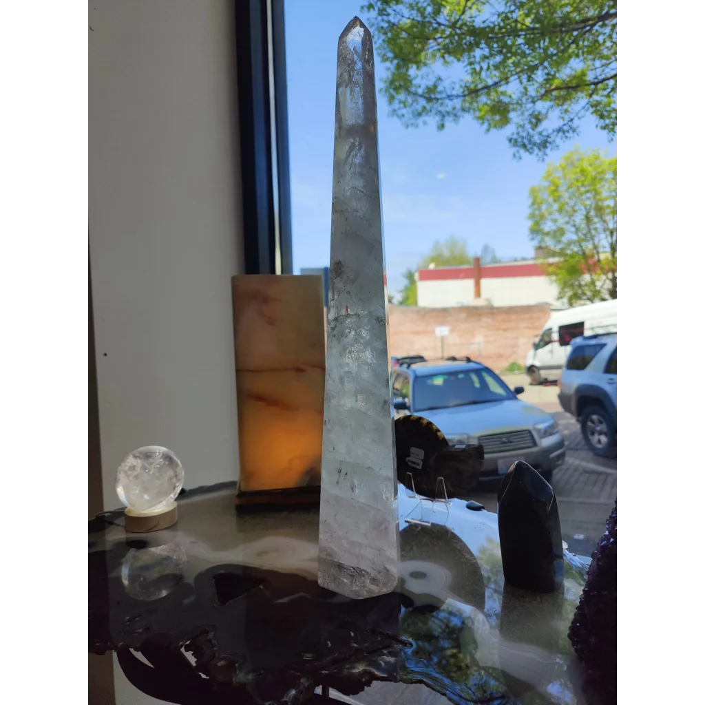 A clear quartz polished obelisk sculpture 20.5 inches tall with natural inclusions and a pointed top on a table