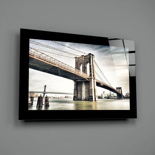 Black-framed glass wall art mounted on gray wall displaying Brooklyn Bridge spanning water with Manhattan skyline and cloudy sky.