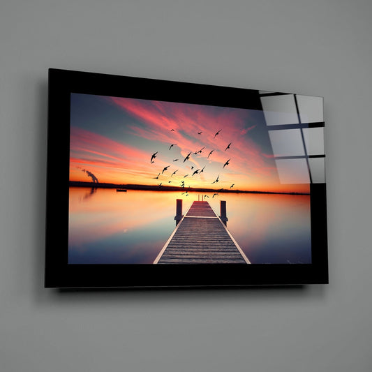 Wall-mounted glass art panel displaying a wooden pier extending into calm water at sunset with flying birds and pink-orange sky.