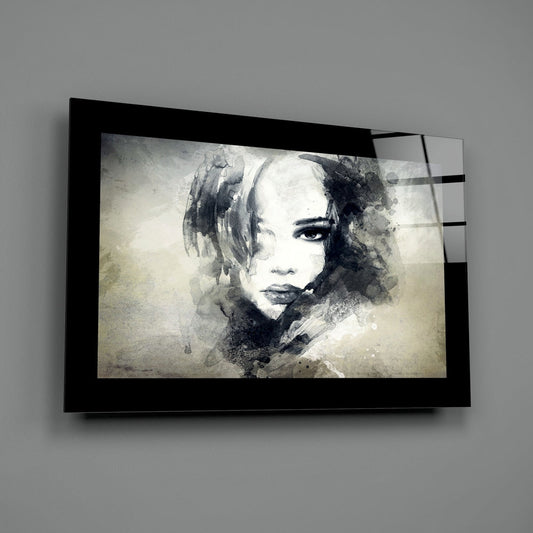 Black-framed glass wall art mounted on gray wall displaying monochromatic watercolor portrait of woman's face with ink splashes.