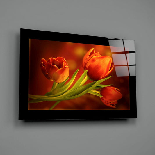 Wall-mounted glass art displaying three red-orange tulips with green stems against a warm amber background, shown at an angle on gray wall.