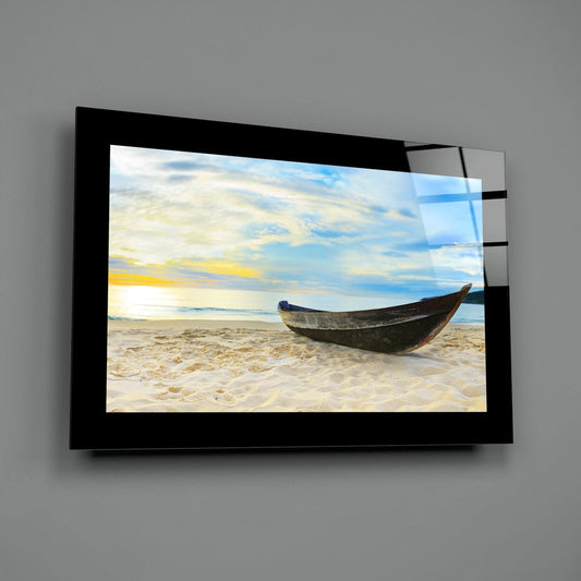 Black-framed glass wall art displaying wooden boat on sandy beach at sunset, mounted on gray wall.