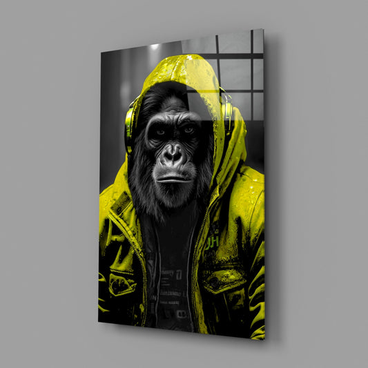 A chimpanzee wearing a vibrant yellow rain jacket and headphones stares directly at the camera against a monochromatic urban background, displayed as a glass wall art panel.