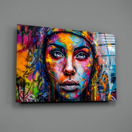 Colorful street art glass wall panel featuring a woman's face covered in vibrant paint splatters and graffiti marks, displayed at an angle against a gray wall.