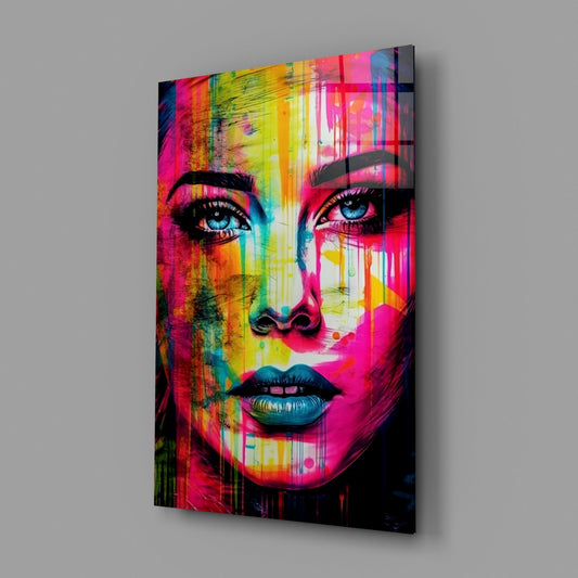 Angled view of a glass wall art panel displaying a vibrant pop art portrait with neon pink, yellow, and cyan colors featuring a woman's face with striking blue eyes.