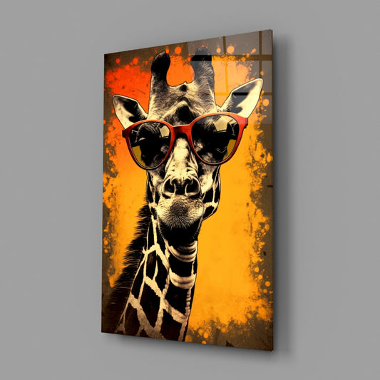 Angled view of a glass wall art panel displaying a stylized giraffe wearing red heart-shaped sunglasses against an orange and yellow gradient background with splatter effects.