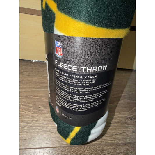 Rolled Green Bay Packers fleece throw with yellow stripe and black care label visible against wooden floor.