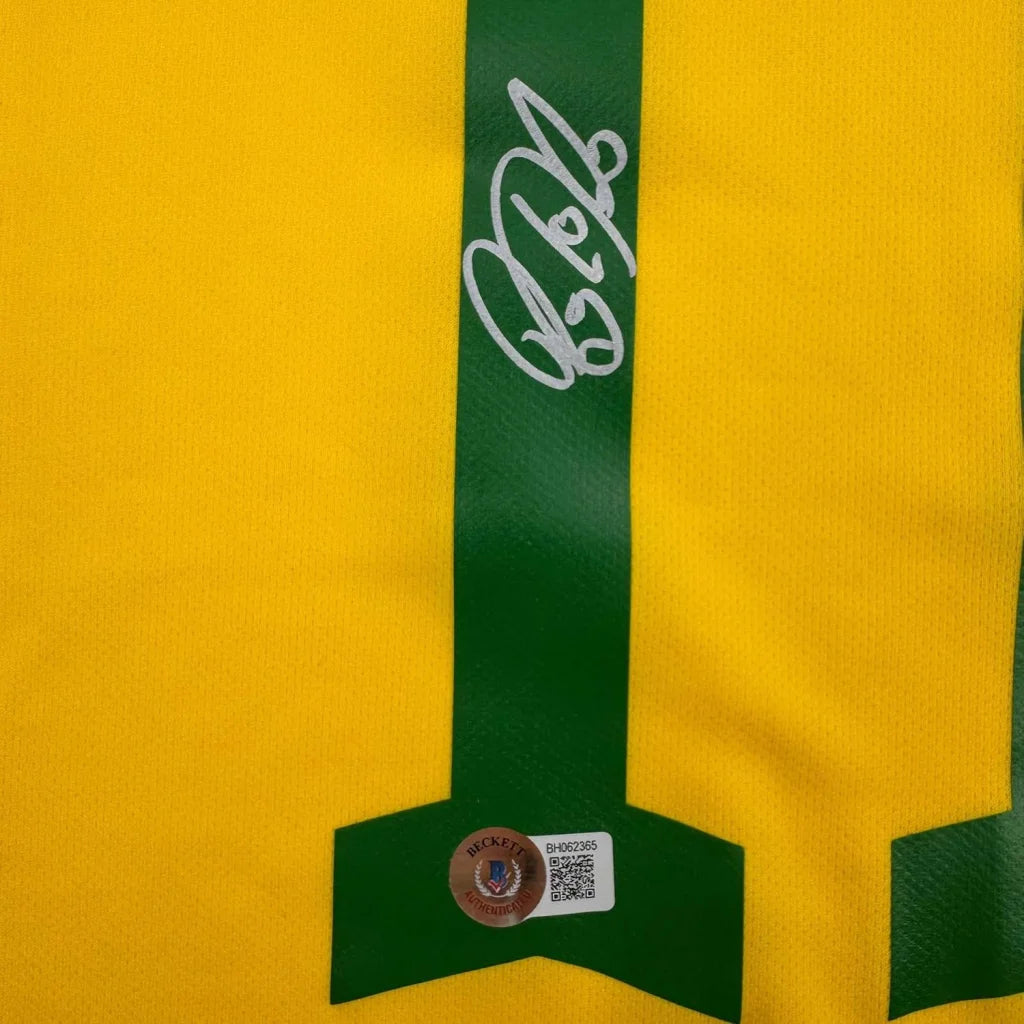 Framed autographed yellow and green Brazil soccer jersey with white signature on green vertical stripe by Raphinha