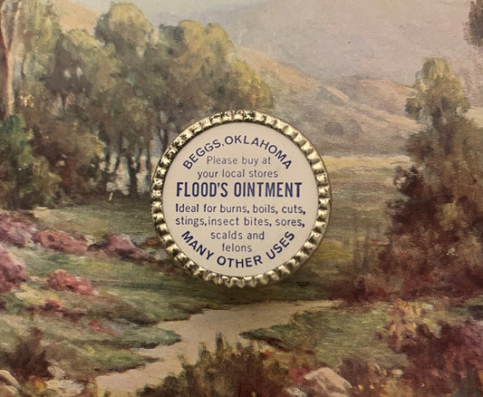 Vintage Flood's Ointment tin lid with gold-ridged rim displayed on a painted landscape background showing trees and meadow.