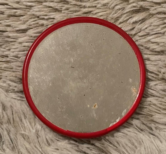 Vintage red-rimmed pocket mirror with cream-colored face on textured beige fabric background, showing wear and patina.