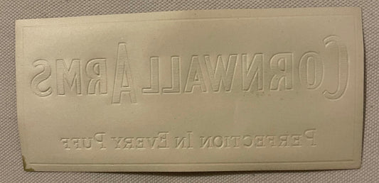 Cream-colored embossed cigar label with "CORNWALL ARMS" text and reversed lettering below, photographed flat against beige fa