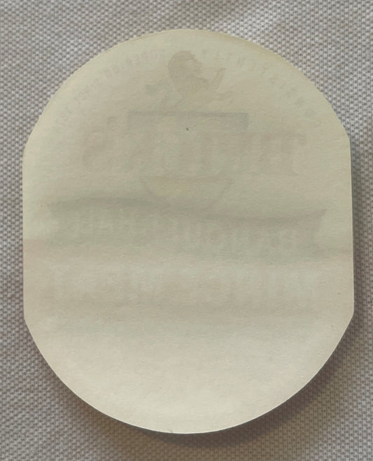 Cream-colored oval tin label with faint embossed text, photographed flat against gray textured background