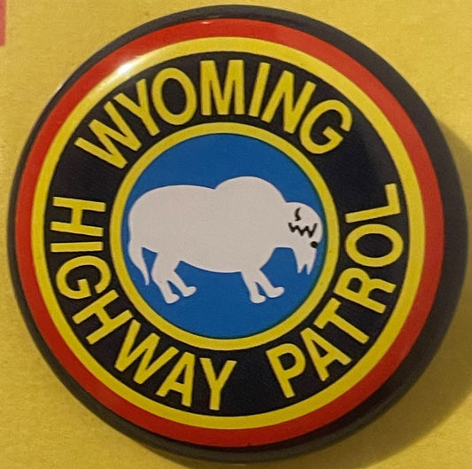 Circular tin litho badge with white buffalo on blue center, yellow text reading "Wyoming Highway Patrol" on black band, red a