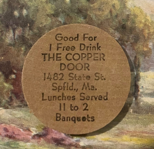 Wooden nickel token with embossed text reading "Good For 1 Free Drink THE COPPER DOOR" and address details, photographed agai