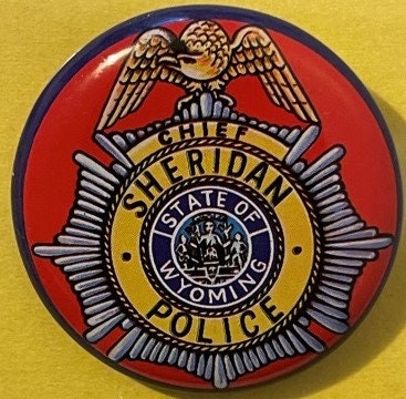 Circular red and gold police badge with eagle emblem and "Chief Sheridan State of Wyoming Police" text on yellow background