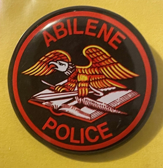 Circular red and black pin-back button with winged eagle holding open book, "ABILENE POLICE" text, displayed against yellow b
