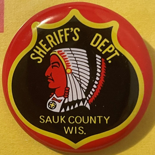 Circular tin litho badge with red rim and black shield featuring a Native American head profile in red, "SHERIFF'S DEPT." and
