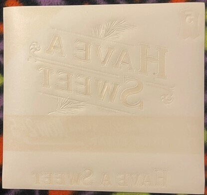 Embossed cream-colored cigar label with "Have A Sweet" text and decorative leaf designs, shown at an angle against colorful f