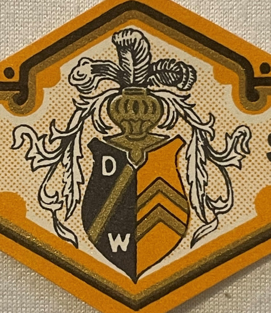 Antique heraldic crest label with orange and black shield, ornate leaf flourishes, and letters D and W on beige background wi