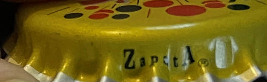 Close-up of yellow vintage bottle cap with red and black dots pattern and partial text visible on white surface