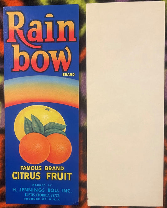 Vintage Rainbow Brand citrus crate label with two oranges, yellow sun, and gradient sky on blue background, displayed flat ne