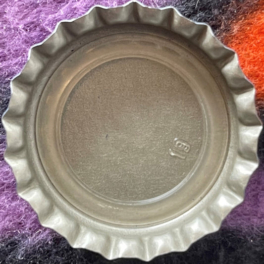 Vintage Royal Crown Cola bottle cap with ridged edges on colorful fabric background, embossed mark visible on tan metal surfa