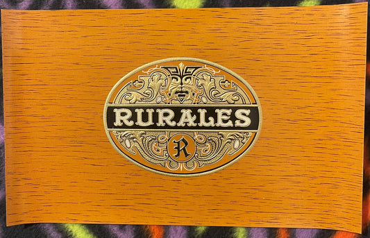 Ornate oval embossed metal label reading "RURALES" with decorative scrollwork on wood-grain cigar box lid, centered view.