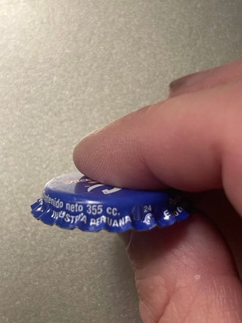 Close-up of blue RC Cola bottle cap held between fingertip and thumb, showing embossed text and ridged crown edge details.