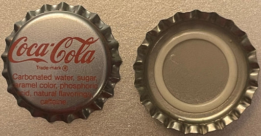 Two vintage Coca-Cola bottle caps shown front and back against a tan textured surface; front cap displays red Coca-Cola scrip